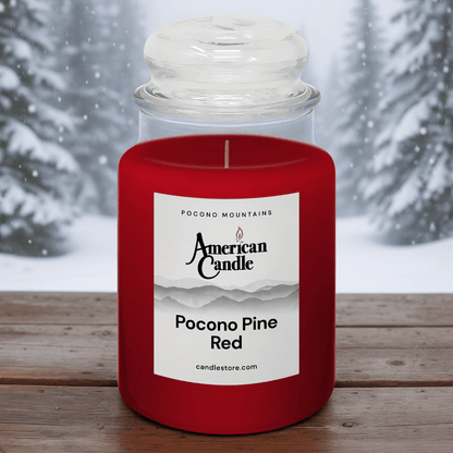 American Candle Pocono Pine Red Scented Jar Candle on a Wooden Table in front of the Wintery Pine Trees